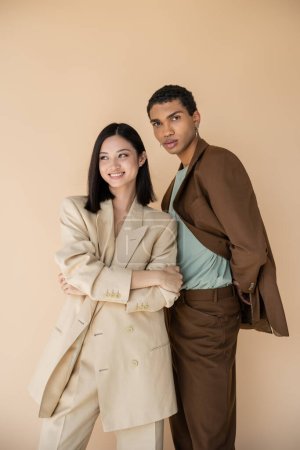 cheerful asian woman posing with folded arms near african american man in brown suit isolated on beige