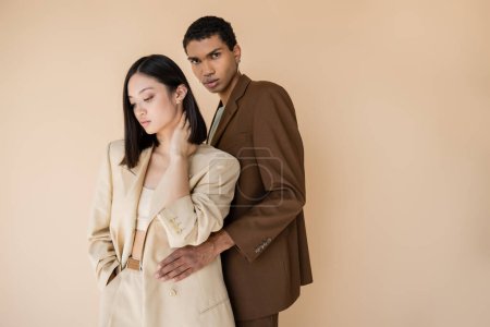 young african american man in brown blazer embracing brunette asian woman and looking at camera isolated on beige