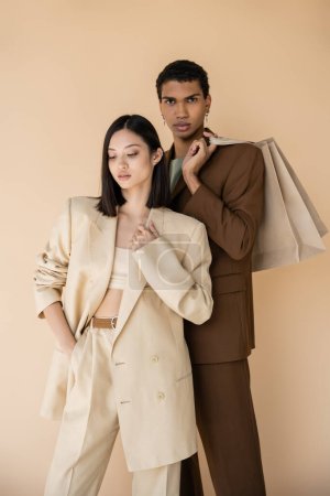 Afrikanisch-amerikanischer Mann mit Einkaufstaschen blickt in die Kamera nahe asiatische Frau in Strumpfhosen isoliert auf beige