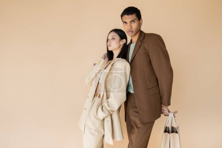 Téléchargez les photos : Afro-américain en costume marron posant avec des sacs à provisions près modèle asiatique à la mode isolé sur beige - en image libre de droit