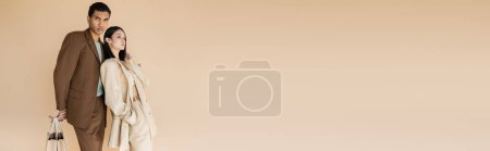 Téléchargez les photos : Afro-américain homme avec des sacs à provisions regardant caméra près élégant asiatique femme avec la main dans la poche isolé sur beige, bannière - en image libre de droit