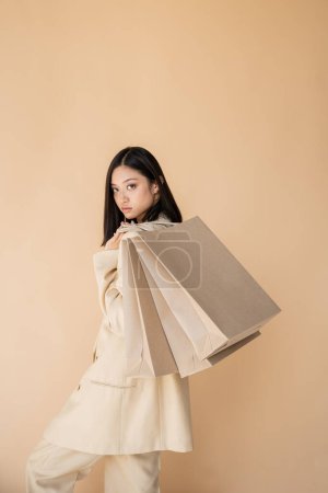 Téléchargez les photos : Jeune et à la mode asiatique femme avec des sacs à provisions regardant caméra isolé sur beige - en image libre de droit