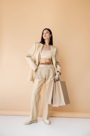 full length of asian woman with shopping bags posing in stylish ivory pantsuit on beige background