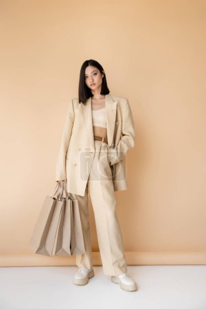 full length of brunette asian woman in stylish pantsuit standing with shopping bags on beige background