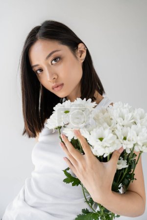 Foto de Morena mujer asiática con piel perfecta y maquillaje natural mirando a la cámara cerca de flores blancas aisladas en gris - Imagen libre de derechos