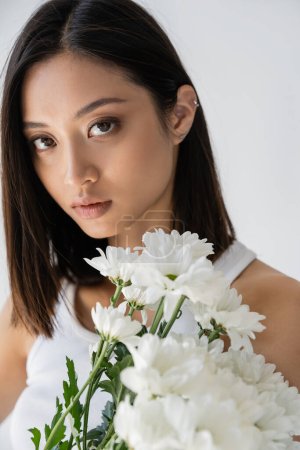Téléchargez les photos : Portrait de femme asiatique sensuelle aux cheveux bruns et maquillage naturel près de fleurs blanches isolées sur gris - en image libre de droit