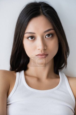 Porträt einer brünetten asiatischen Frau in weißem Tank-Top, die isoliert auf grau in die Kamera blickt