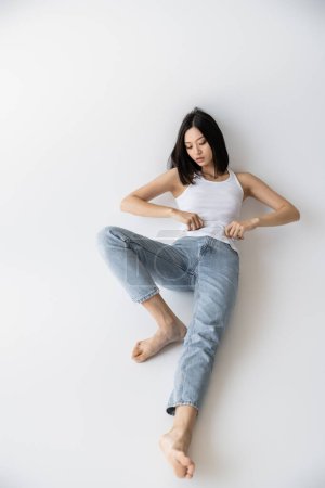 full length of barefoot asian woman in white tank top and jeans lying on grey background