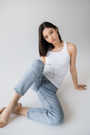pretty asian woman in white tank top and jeans looking at camera while sitting on grey background
