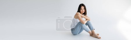 full length of slender asian model in blue jeans and white tank top sitting on grey background, banner