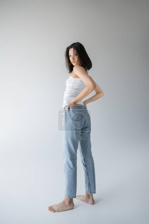 full length of barefoot asian woman in white tank top and blue jeans holding hands in back pockets on grey background
