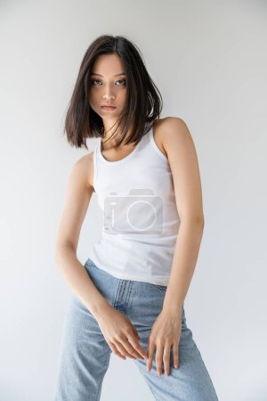 Téléchargez les photos : Brunette asiatique femme en blanc débardeur et bleu jeans regarder caméra isolé sur gris - en image libre de droit