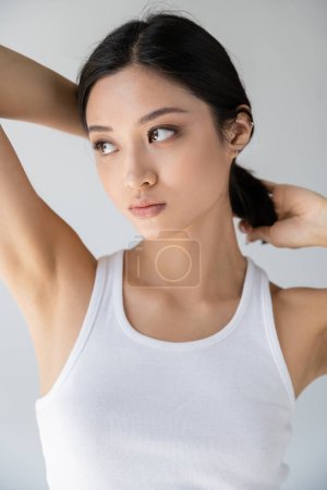 pretty asian woman in white tank top touching brunette hair and looking away isolated on grey