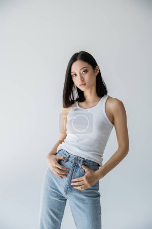modelo asiático joven y delgado posando en pantalones vaqueros azules y camiseta blanca aislada en gris
