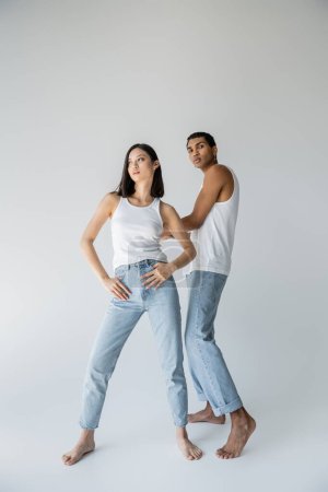 full length of stylish asian woman posing with thumbs in pockets of jeans near barefoot african american man on grey background