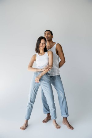 Téléchargez les photos : Longueur totale de pieds nus et mince couple multiethnique en débardeurs et jeans sur fond gris - en image libre de droit