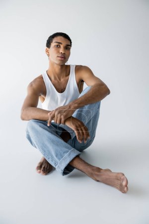 Téléchargez les photos : Homme afro-américain pieds nus en jeans bleus assis avec les jambes croisées et regardant la caméra sur fond gris - en image libre de droit