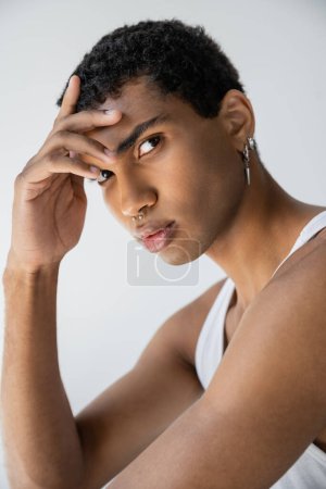 retrato de hombre afroamericano con piercing plateado sosteniendo la mano cerca de la frente y mirando a la cámara aislada en gris