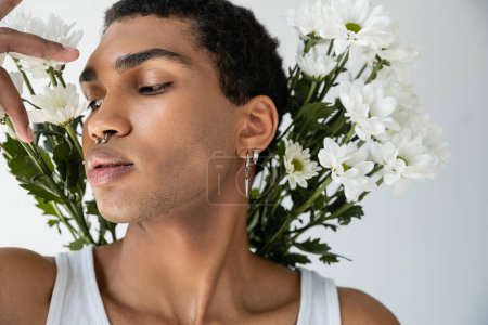 Foto de Retrato de hombre afroamericano con piercing de plata posando cerca de flores frescas blancas aisladas en gris - Imagen libre de derechos