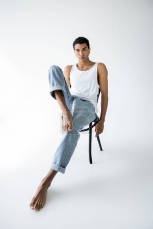 full length of barefoot african american man in blue jeans and white tank top sitting on chair on grey background