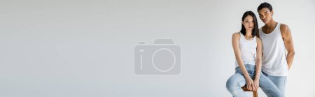 Téléchargez les photos : Brunette asiatique femme en blanc débardeur et jeans assis sur tabouret près afro-américain gars isolé sur gris, bannière - en image libre de droit