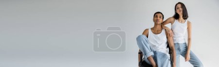 sinnliche asiatische Frau in Jeans und Tank-Top schaut in die Kamera in der Nähe afrikanisch-amerikanischer Kerl sitzt auf Stuhl isoliert auf grau, Banner