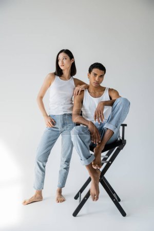 Téléchargez les photos : Pleine longueur de pieds nus asiatique femme en débardeur et jeans regarder loin près afro-américain gars assis sur chaise sur fond gris - en image libre de droit