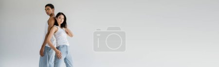 Téléchargez les photos : Élégant couple multiethnique en débardeurs blancs et jeans posant dos à dos et regardant la caméra isolée sur gris, bannière - en image libre de droit