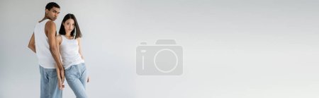 Téléchargez les photos : Jeune couple multiethnique en débardeurs blancs et jeans bleus regardant caméra isolée sur gris, bannière - en image libre de droit