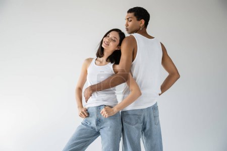 african american man in white tank top and jeans embracing pleased asian woman smiling with closed eyes isolated on grey