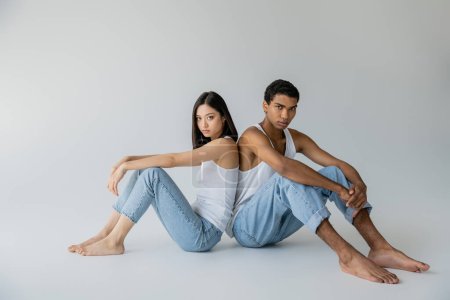 Téléchargez les photos : Longueur totale de pieds nus et élégant couple interracial en jeans et débardeurs assis dos à dos sur fond gris - en image libre de droit