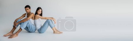 full length of barefoot multiethnic couple in tank tops and jeans sitting back to back on grey background, banner