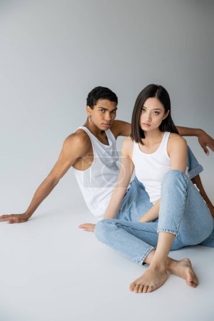 Téléchargez les photos : Couple interracial à la mode en jeans et débardeurs assis et regardant la caméra sur fond gris - en image libre de droit
