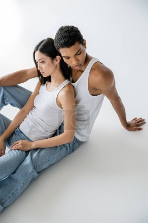 high angle view of young interracial couple in white tank tops and jeans sitting on grey background