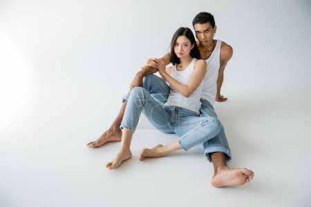 Téléchargez les photos : Pleine longueur de pieds nus couple multiethnique assis en jeans bleus et débardeurs blancs sur fond gris - en image libre de droit