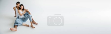 Téléchargez les photos : Longueur totale de couple interracial élégant en jean bleu assis sur fond gris, bannière - en image libre de droit