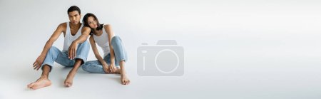 full length of slim multiethnic couple in blue jeans and white tank tops sitting on grey background, banner