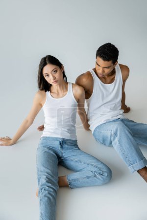 Photo for Slim asian woman in blue jeans and white tank top looking at camera near african american guy on grey background - Royalty Free Image