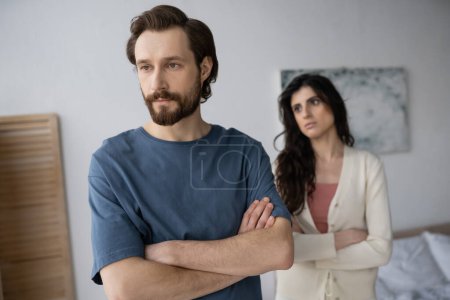 Sad man crossing arms near blurred girlfriend in bedroom at home 