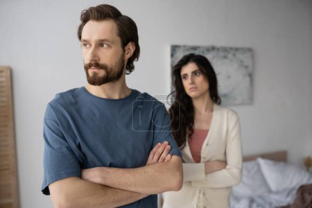 Angry man crossing arms near blurred girlfriend in bedroom 