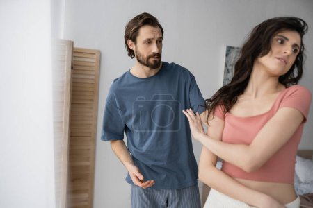 Bearded man in pajama talking to tensed girlfriend in bedroom at home 