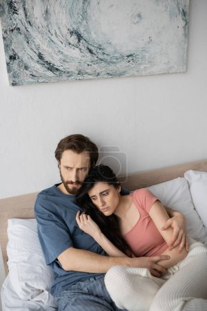 Dissatisfied man in pajama hugging asexual girlfriend on bed at home 