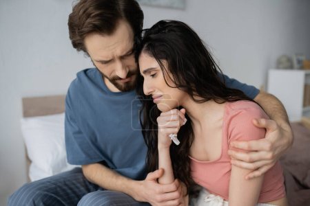 Empathetic man in pajama calming crying girlfriend with napkin in bedroom 