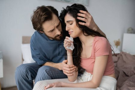 Téléchargez les photos : Homme attentionné en pyjama étreignant pleurer et bouleversé petite amie sur le lit à la maison - en image libre de droit