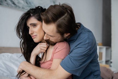 Bärtiger Mann umarmt traurige Freundin mit Serviette im verschwommenen Schlafzimmer 