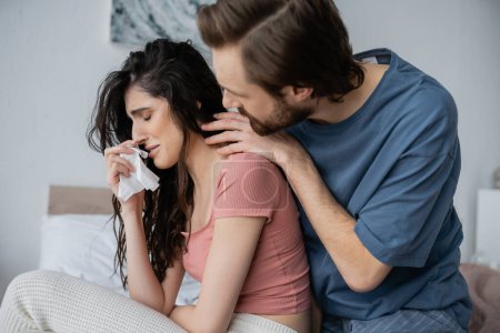 Caring man in pajama hugging crying girlfriend with napkin in bedroom 