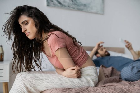 Upset woman in pajama feeling bad and sitting near blurred boyfriend with smartphone in bedroom 