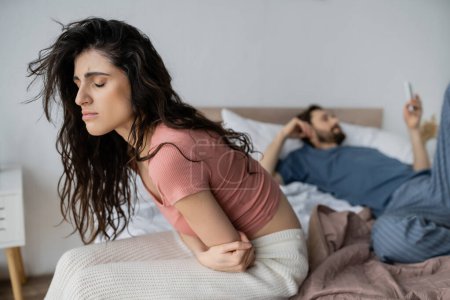 Enttäuschte brünette Frau fühlt sich schlecht, während sie neben verschwommenem Freund mit Smartphone auf dem Bett zu Hause sitzt 