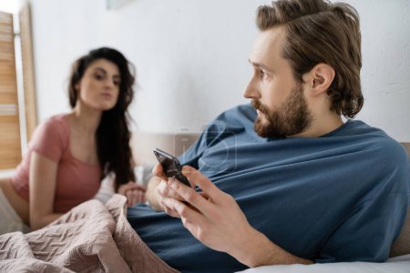 Bearded man using smartphone near blurred jealous girlfriend in bedroom 
