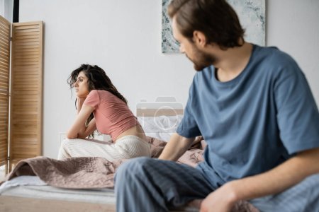 Indifferent woman in pajama sitting on bed near blurred boyfriend at home 
